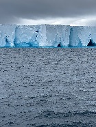 Cloud covered sky hovers just over a plateau of ice seen from the a position across a choppy sea